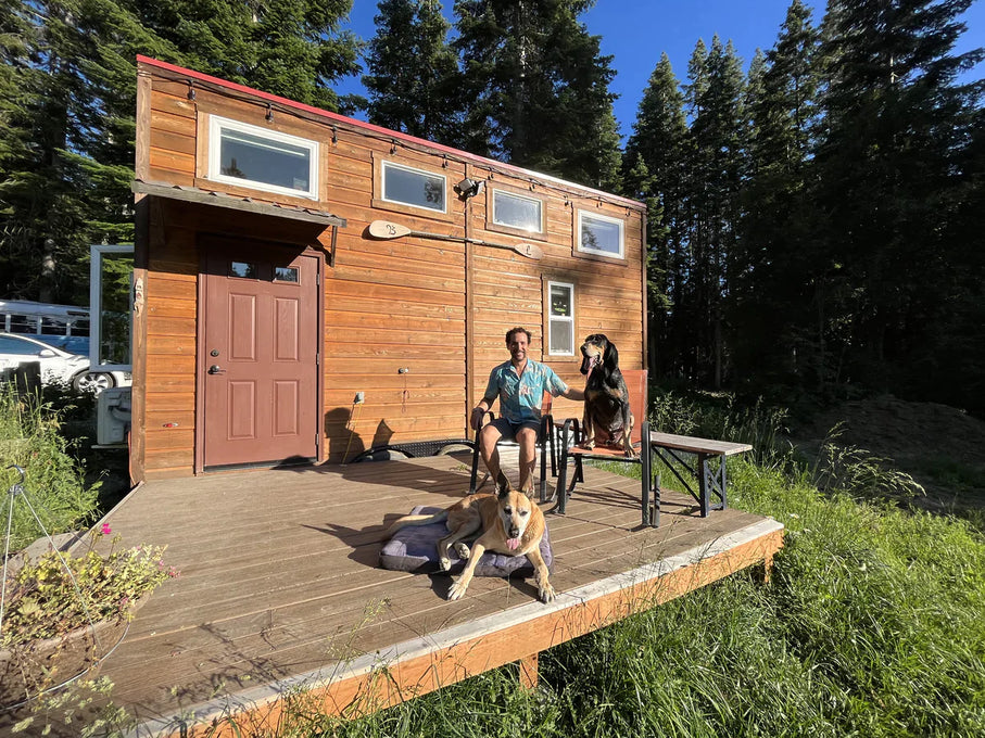 Man sits with two big dogs on porch of tiny house.